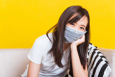 Portrait Asian of beautiful young woman sitting on sofa wearing medical face mask protective during Coronavirus studio shot isolated on yellow backgroundの写真素材