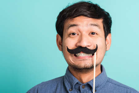 Portrait Asian happy handsome man posing he holding a funny mustache card or vintage fake moustaches on his mouth, studio shot isolated on blue background, Fathers day, November day conceptの写真素材
