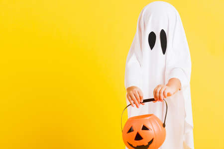 Funny Halloween Kid Concept, little cute child with white dressed costume halloween ghost scary he holding orange pumpkin ghost on hand, studio shot yellow on white backgroundの写真素材