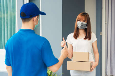 Asian delivery express courier young man giving parcel boxes to woman customer receiving both protective face mask and show thumbs up finger for good support sign, under curfew pandemic coronavirusの写真素材