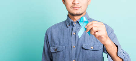 Asian portrait happy handsome man posing he holding light blue ribbon for supporting people living and illness, studio shot isolated on blue background, Prostate Cancer Awareness in November conceptの写真素材