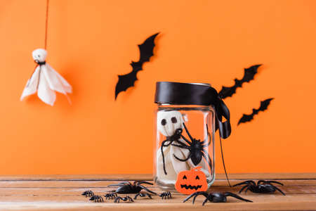 Funny Halloween day decoration party, Baby white ghost crafts scary face in jar glass on wood table, studio shot isolated on orange background have spider and bats, Happy holiday DIY conceptの写真素材