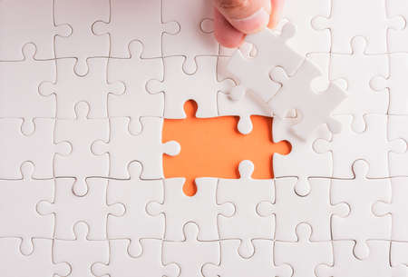 Top view flat lay of hand-holding last piece white paper jigsaw puzzle game last pieces put to place for solve problem complete mission, studio shot on an orange background, quiz calculation conceptの写真素材
