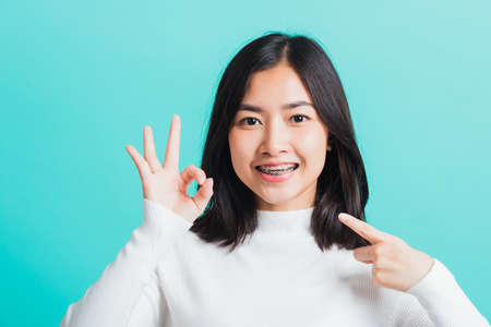 Asian teen beautiful young woman smile have dental braces on teeth laughing she showing gesturing ok sign with fingers, isolated on a blue background, Medicine and dentistry conceptの写真素材