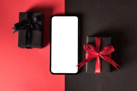 Black Friday sale shopping concept, Top view of gift box wrapped in black paper and black bow ribbon and modern mobile smartphone, studio shot on red and dark backgroundの写真素材