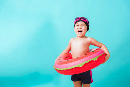 Summer vacation concept, Portrait Asian happy cute little child boy wear goggles and swimsuit hold watermelon inflatable ring, Kid having fun on summer vacation, studio shot isolated blue backgroundの写真素材