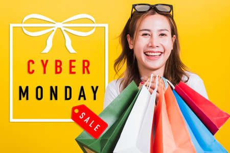 Asian happy portrait beautiful young woman smiling stand with sunglasses excited holding shopping bags multi color looking camera with Cyber Monday text in gift box on side isolated yellow backgroundの写真素材