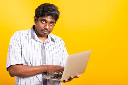 Asian happy portrait young black man smiling standing wear shirt holding and typing laptop computer he looking to camera isolated, studio shot yellow background with copy spaceの写真素材