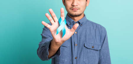 Asian portrait happy handsome man posing he holding light blue ribbon for supporting people living and illness, studio shot isolated on blue background, Prostate Cancer Awareness in November conceptの写真素材
