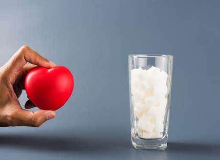 Hand hold heart near glass full of white sugar cube sweet food ingredient studio shot isolated on gray background, health high blood risk of diabetes and calorie intake concept and unhealthy drinkの写真素材