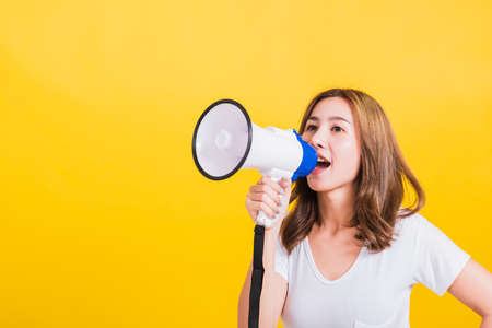 Asian Thai happy portrait beautiful cute young woman stand to make announcement message shouting screaming in megaphone looking to side away, studio shot isolated on yellow background with copy spaceの写真素材