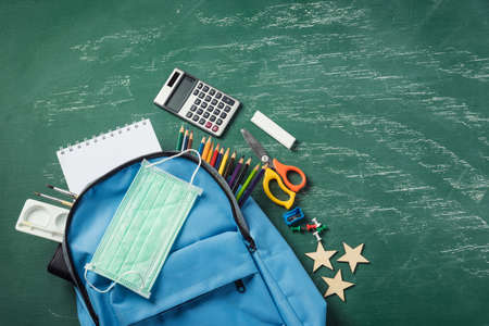 Top view of stylish blue school bag backpack at a green chalkboard with face mask protection and...