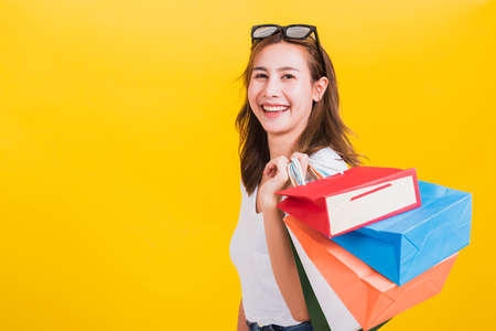 Asian Thai portrait happy beautiful cute young woman smiling stand with sunglasses excited holding shopping bags multi color looking camera, studio shot isolated yellow background with copy spaceの写真素材