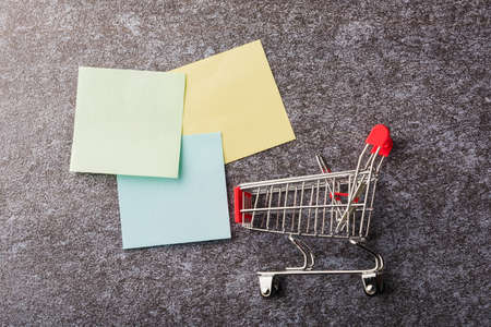 Above blank paper stick note list yellow and green with the shopping cart on concrete background,...