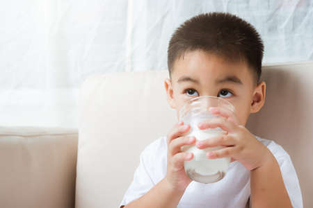 Close up of happy Asian little cute child boy hand holding milk glass he drinking white milk during sitting on the sofa at home after lunch. Daily life health care Medicine foodの写真素材