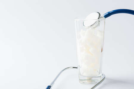 A glass full of white sugar cube sweet food ingredient and doctor stethoscope, studio shot isolated white background, health high blood risk of diabetes and calorie intake concept and unhealthy drinkの写真素材