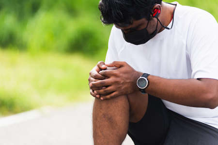 Close up Asian young sport runner black man wear watch sitting hands joint hold knee pain during the run while running at the outdoor street health park, healthy exercise injury from workout conceptの写真素材