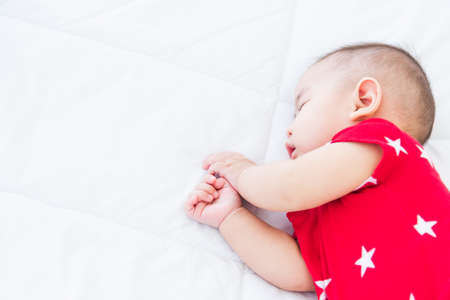 Portrait of beautiful Asian newborn little baby sleeping on white bed at home, Happy baby wears a red shirt relaxing pillow in the room, Family morning at homeの写真素材