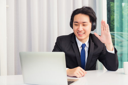 Work from home, Asian young businessman smile wearing headphones and suit video conference call or video call on desk and raise your hand to say hello greet colleague at home officeの写真素材