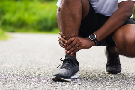 Close up Asian young sport runner black man wear watch hands joint hold leg pain because of twisted ankle broken while running at the outdoor street health park, healthy exercise Injury from workoutの写真素材