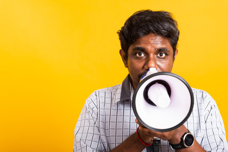 Asian happy portrait young black woman standing to make announcement message shouting screaming in megaphone, studio shot isolated on yellow background with copy spaceの写真素材