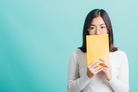 Young beautiful Asian woman hiding behind an open book, Portrait female in glasses is holding and reading a book, studio shot isolated on a blue background, Education conceptの写真素材