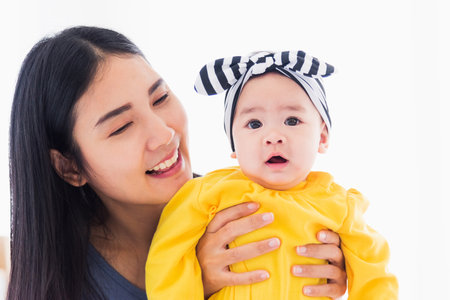 Portrait of beautiful young Asian mother playing and smiling together with his newborn little baby at home, Parent mom and little kid relaxing in the bedroom, Family having fun togetherの写真素材