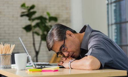 Asian businessman tired overworked he sleeping over a laptop computer on the desk. senior man with eyeglasses lying asleep on table at his working placeの写真素材