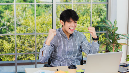 Yes! Happy excited Asian man raising his arm up to celebrate celebrating success. Young businessman using laptop computer at office desk he glad to receive good winner profits from the jobの写真素材