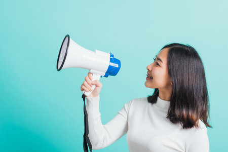 Beautiful Asian woman smile she holding megaphone making the announcement, female excited cheerful announce good news isolated on a blue backgroundの写真素材