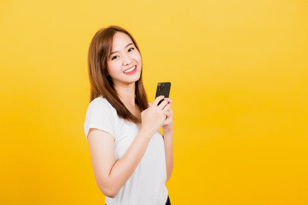 Asian happy portrait beautiful cute young woman teen smiling standing wear t-shirt using smart mobile phone looking to camera isolated, studio shot on yellow background with copy spaceの写真素材