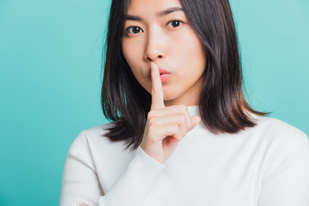 Young beautiful Asian woman holding index finger on her mouth lips, Portrait female hush silence, studio shot isolated on a blue background, Gesture of shhhの写真素材