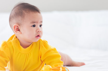 Portrait of beautiful young Asian newborn little baby prone on the bed at home, Happy baby smile wears a yellow shirt relaxing in the room, Family morning at homeの写真素材
