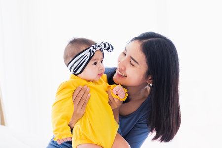 Portrait of beautiful young Asian mother playing and smiling together with his newborn little baby at home, Parent mom and little kid relaxing in the bedroom, Family having fun togetherの写真素材