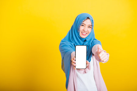 Asian Muslim Arab woman Islam wear veil hijab funny smile she showing blank screen smart mobile phone and point screen, studio shot isolated on yellow backgroundの写真素材
