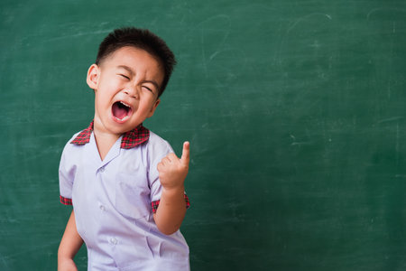 Back to School. Happy Asian funny cute little child boy from kindergarten in student uniform smiling point finger to side away space on green school blackboard, First time to school education conceptの写真素材