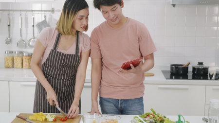 Happy Asian beautiful young family couple husband and wife cooking vegetable salad in kitchen together at home. The man and woman according online cooking class to recipe on laptop computerの写真素材