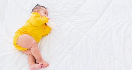 Asian beautiful little baby girl wearing a yellow dress eat milk sleeping feeding lying on the white bed, infant holding a bottle of milk, baby food conceptの写真素材