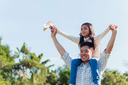 Happy Asian young family father and carrying an excited girl on shoulders having fun and enjoying outdoor lifestyle together playing aircraft toy on sunny summer day, Father's day conceptの写真素材