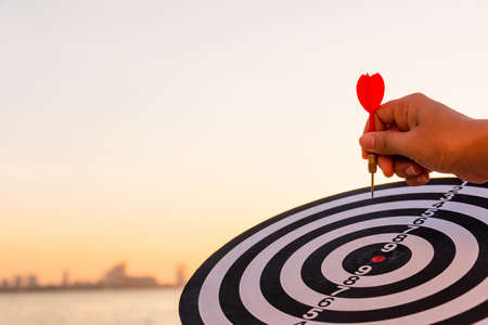 Hand hold of dart arrow on bullseye (bull's-eye) dartboard is the target of purpose challenge business at sunset, expert marketing strategy target, objective financial and goal successの写真素材