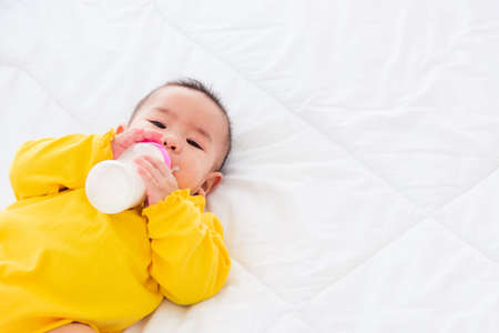 Asian beautiful little baby girl wearing a yellow dress eat milk sleeping feeding lying on the white bed, infant holding a bottle of milk, baby food conceptの写真素材