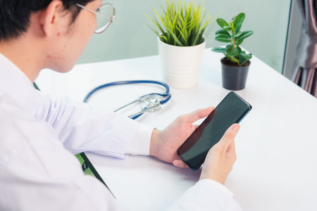 Asian doctor young handsome man smiling using working or holding with digital smart mobile phone at hospital desk office, Technology healthcare medical conceptの写真素材