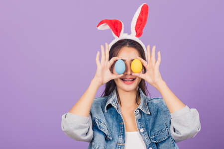 Happy beautiful young woman smiling wearing rabbit ears and denims holding colorful Easter eggs...