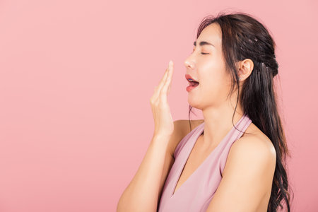 Portrait Asian beautiful young woman emotions tired and sleepy her yawning covering mouth open by hand, studio shot isolated on pink background, Thai female insomnia conceptの写真素材