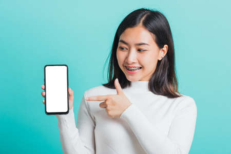 Beautiful Asian woman smile holding a smartphone on hand and pointing finger to the blank screen, female excited cheerful her show mobile phone isolated on a blue background, Technology conceptの写真素材