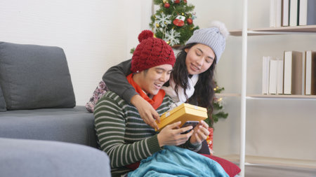 Happy Asian beautiful young family couple husband and wife in home living room, woman hide surprise giving gift box her boyfriend present during play smartphone, winter holidays and valentine conceptの写真素材