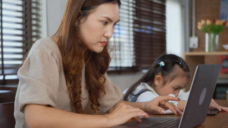 Asian young mother working with laptop computer at home and help daughter doing homework, Single mom freelancer work from home and helping his girl kid teaching homework on deskの写真素材