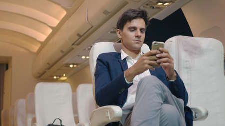 Businessman sitting inside plane watching video on mobile phone during his business trip, Young...