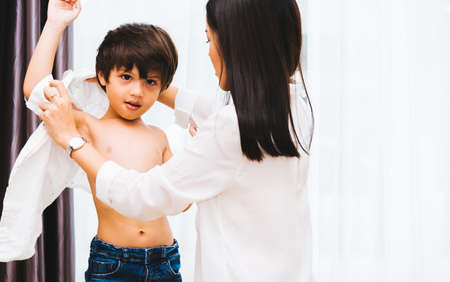Asian happy beautiful mother helping to get dressed clothes on her boy kid in house for ready before...