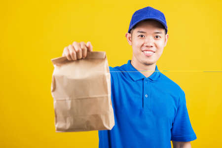 Portrait excited delivery service man smile wearing blue t-shirt and cap uniform hold paper containers for takeaway bag grocery food packet looking to camera, studio shot isolated on yellow backgroundの写真素材